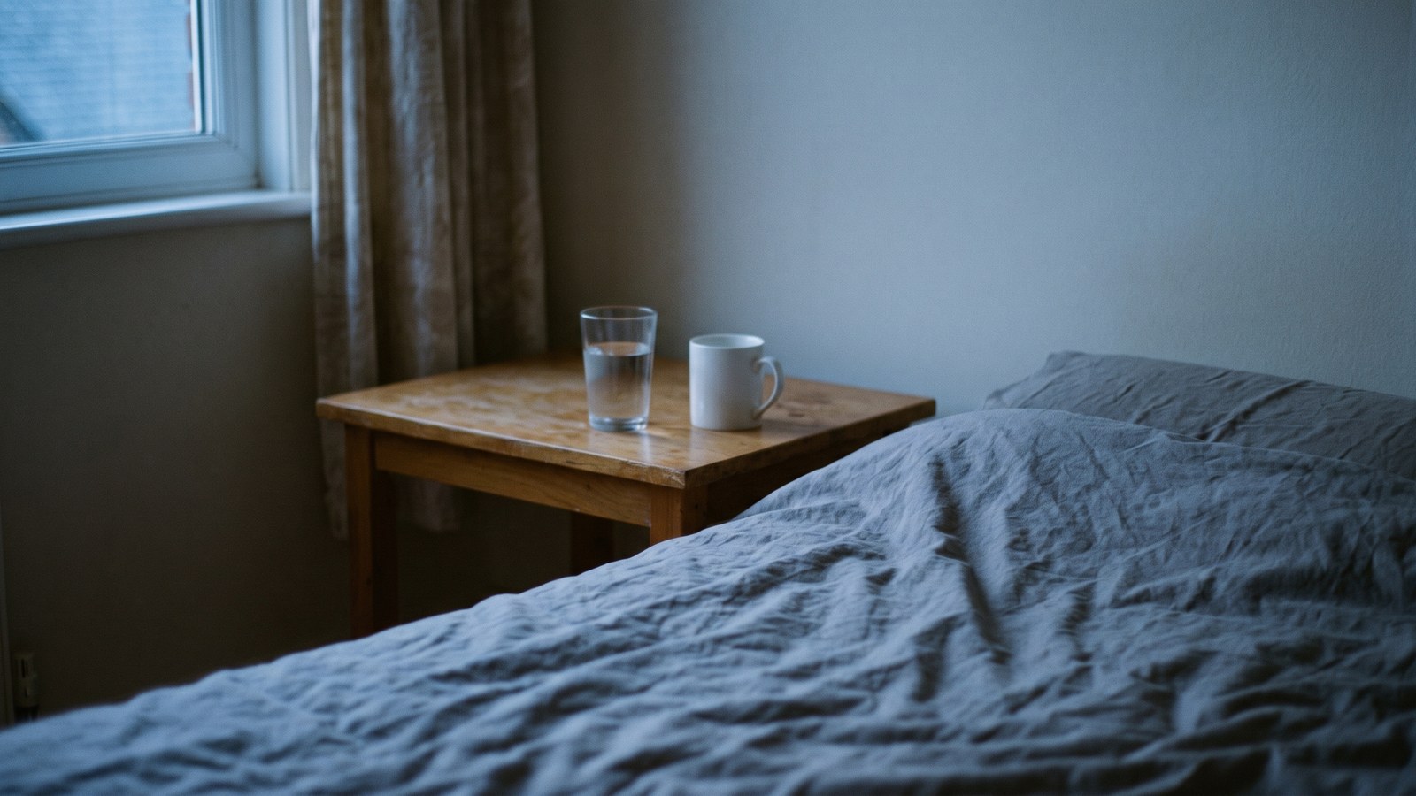 A quiet bedroom at dusk with a glass of water and a plain white mug on a wooden bedside table beside a rumpled grey duvet