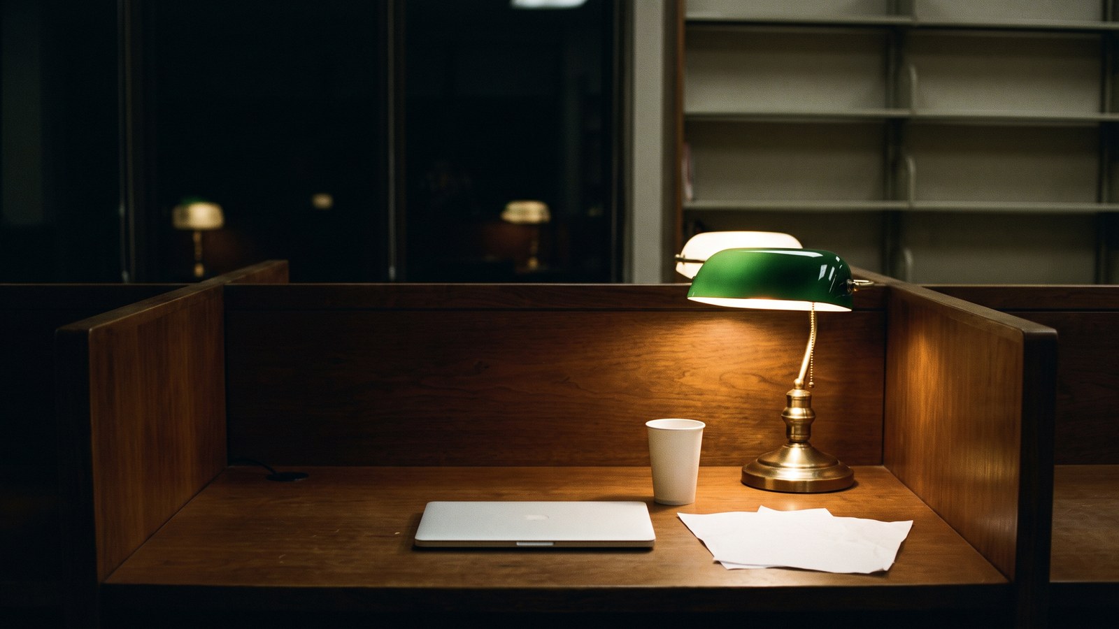 An empty library study carrel at night: a wooden desk with a closed laptop, a plain white paper cup, two blank sheets of paper and a green-shaded desk lamp glowing, with dark windows behind