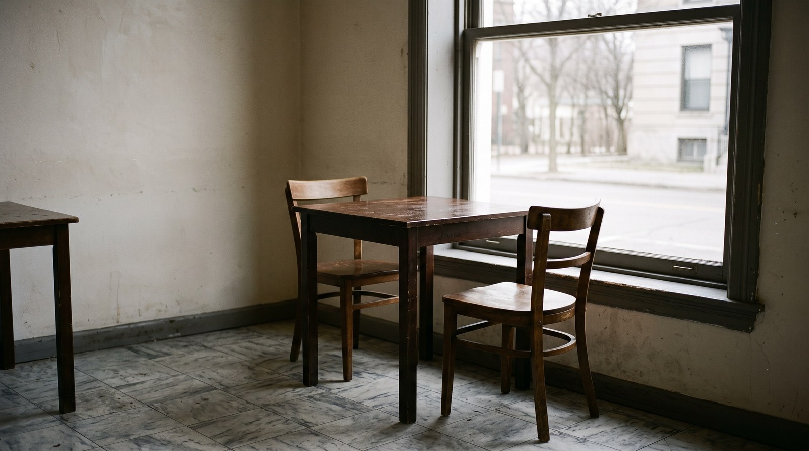 An empty café interior at a quiet hour: one small bare wooden table for two by a large window, two wooden chairs with one pushed in and one turned slightly out, flat grey daylight falling across the tabletop and a worn tiled floor.