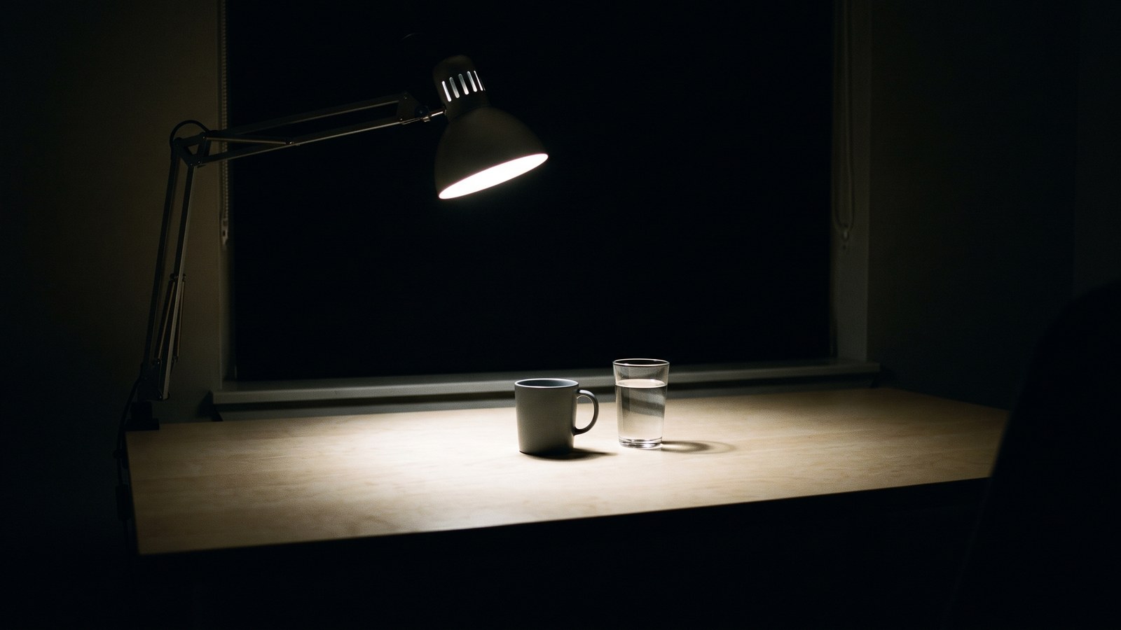 A plain birch desk in a room that is otherwise completely dark, the window behind it solid black. A single aluminium-armed daylight lamp is angled down over it, throwing one intense cold-white pool of light across the desktop. Inside the pool sit a plain unmarked mug and a glass of water. Beyond the edge of the light the room disappears entirely.