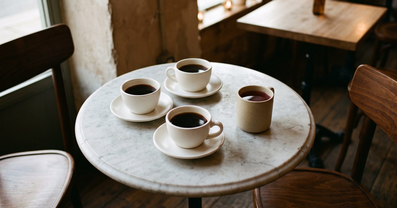 Overhead view of a small round marble cafe table in warm evening light. Three white cups of black coffee on saucers sit clustered together, and one plain oatmeal stoneware mug of dark herbal brew sits slightly apart from them with no saucer. Dark walnut chair backs enter the frame at the edges.