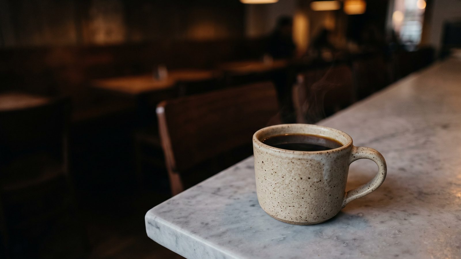 A corner of a cafe's pale grey marble counter in warm evening light, shot low and close. One plain oatmeal stoneware mug of dark, almost black brew sits on the marble with steam curling from it. Behind it the cafe falls away into soft warm out-of-focus darkness lit by a single pendant.
