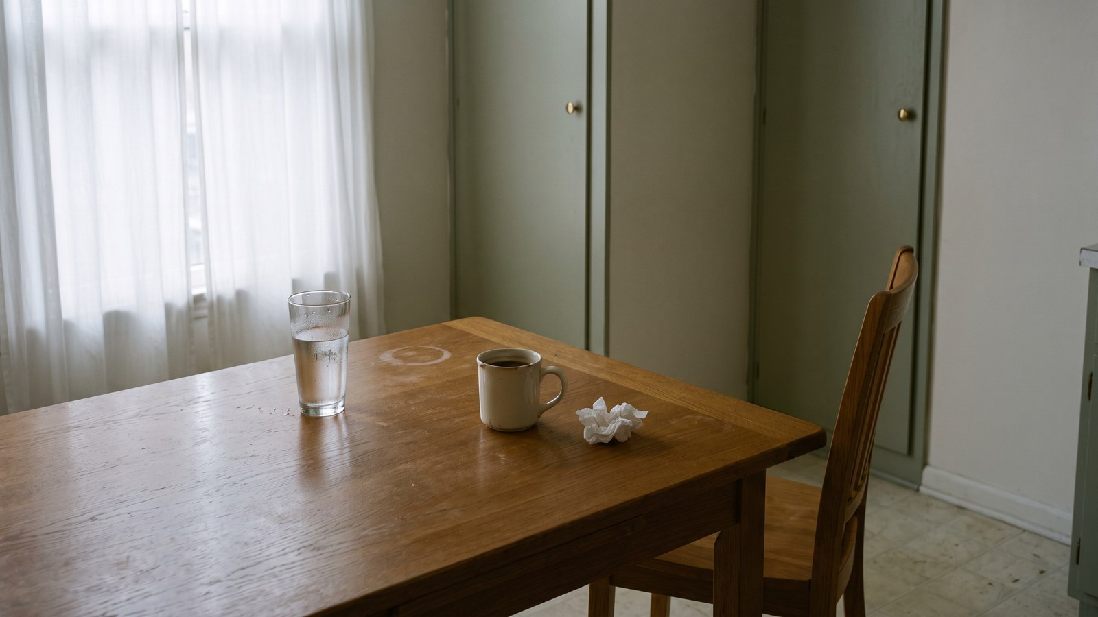 The same dining nook with the white curtains pulled fully closed, the room dim and flat with only a soft grey glow through the fabric. On the oak table: a tall glass of water two-thirds drunk, a cold half-inch of dark drink left in the cream mug, and a crumpled tissue. The chair is pushed right back at an abandoned angle.