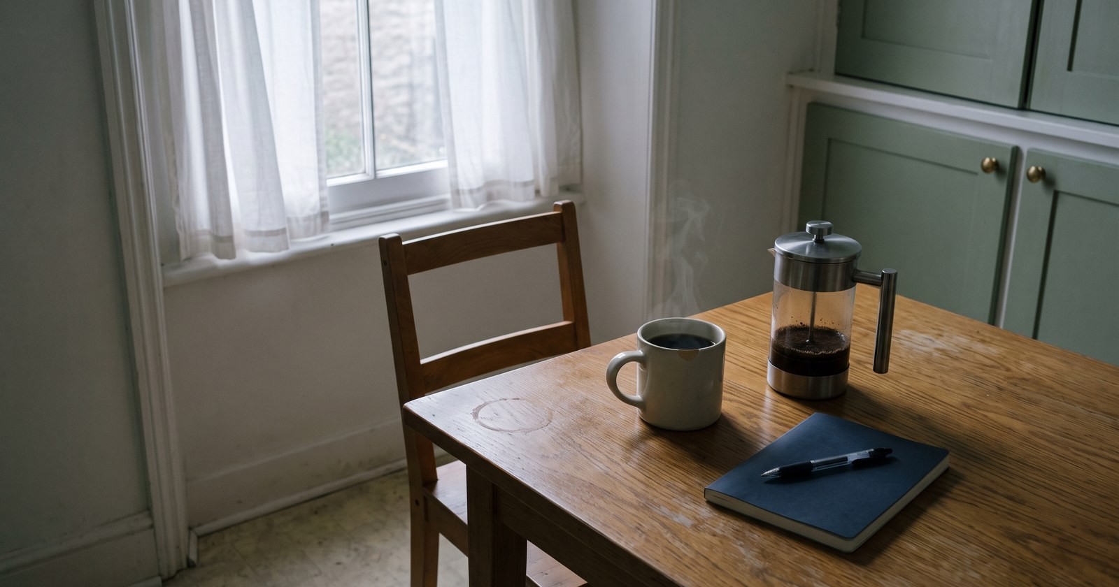 A scratched oak dining table in flat grey pre-dawn light beside a tall curtained window. On it: a chipped cream stoneware mug of almost-black brewed drink faintly steaming, a stainless steel French press with dark grounds settled in the bottom, and a closed navy notebook with a ballpoint pen on top. One wooden chair is pulled out and empty.
