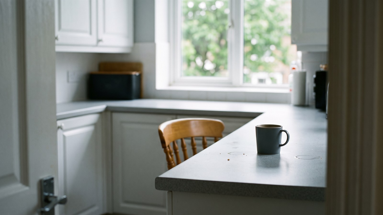 Seen from a doorway into a plain white kitchen in flat, even mid-morning light, green leaves out of focus through the window. Well off to one side of a grey worktop sits a plain unmarked dark mug, already half drunk and no longer steaming, set down casually rather than arranged. Most of the frame is empty worktop and ordinary light.