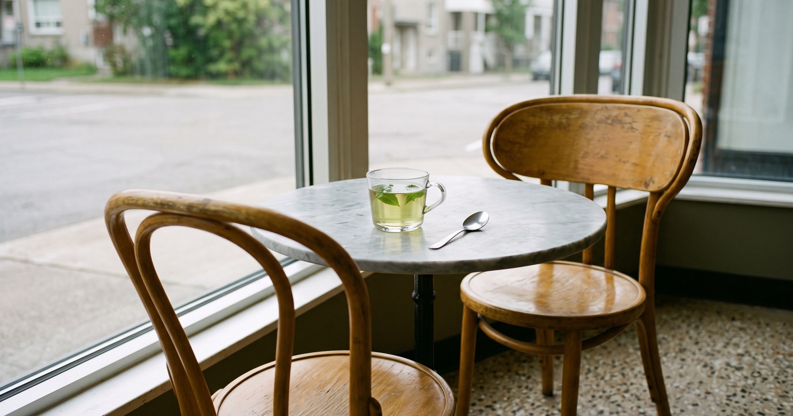A small round marble table by a cafe's plate-glass window in flat, even afternoon light. On it a clear glass cup of pale green mint tea, barely touched and no longer steaming, with a spoon laid beside it. Two honey-toned bentwood chairs; the one opposite is empty and pushed in. Beyond the glass, only out-of-focus street and green trees.