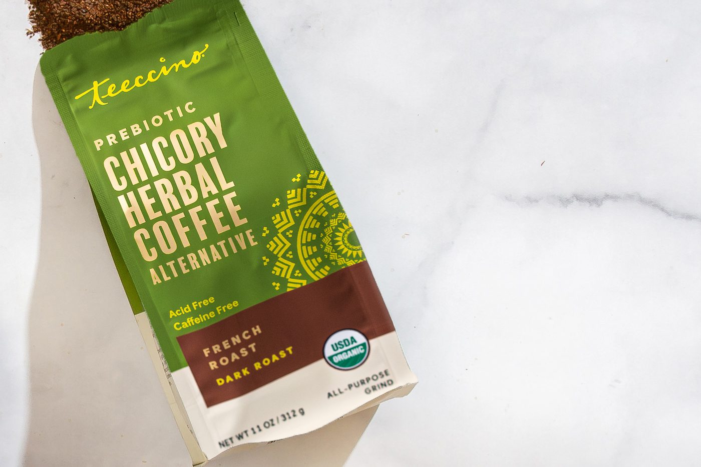 An 11-ounce green foil bag of Teeccino Prebiotic Chicory Herbal Coffee Alternative in French Roast, dark roast, lying on white marble. The front reads acid free and caffeine free and carries a USDA Organic seal and an all-purpose grind note, with a scatter of dark reddish-brown grounds spilling from the open top.