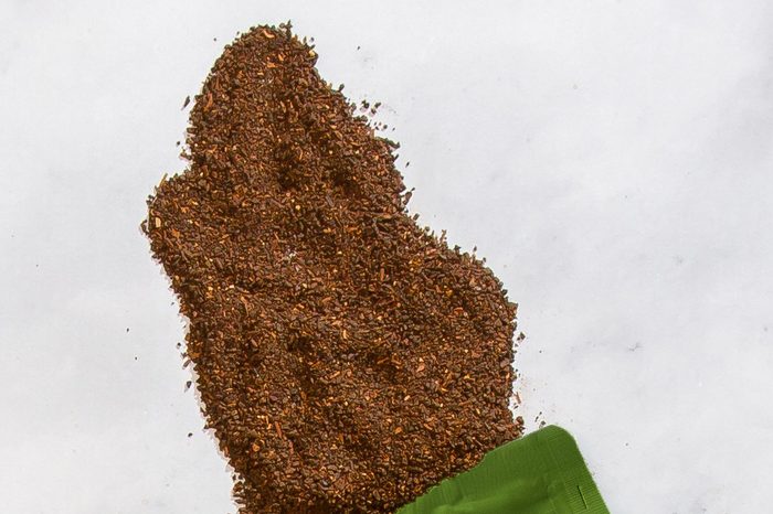 A close view of dry Teeccino French Roast grounds spilled on white marble. The particles are coarse and irregular in size, reddish-brown, and visibly fibrous and woody rather than the even dark powder of ground coffee.